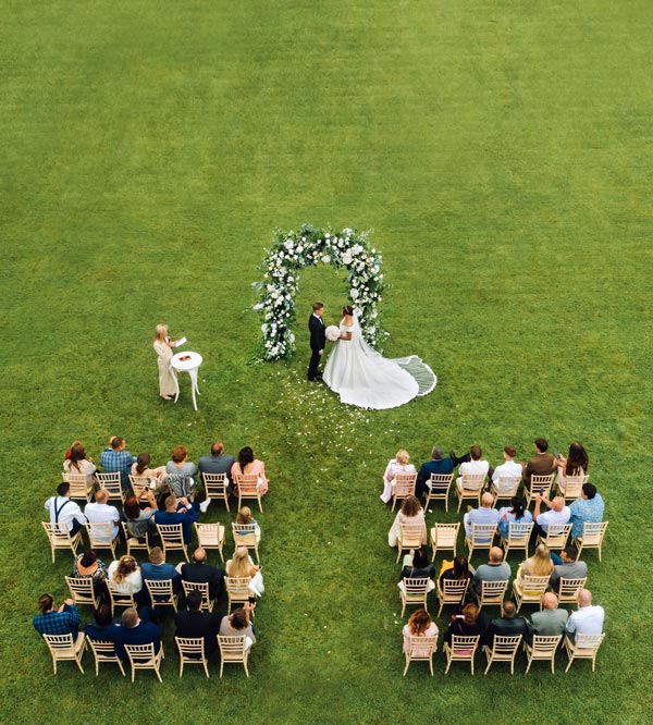 outdoor wedding ceremony with couple standing under floral arch surrounded by guests seated on grass three weddings celebration