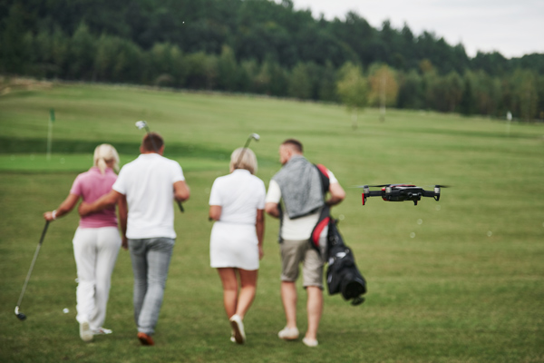 four people walking on golf course with a drone flying above them